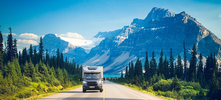 Camping-car profilé étroit Pilote roulant sur une route bordée de sapins, la montagne enneigée et un lac en arrière plan.